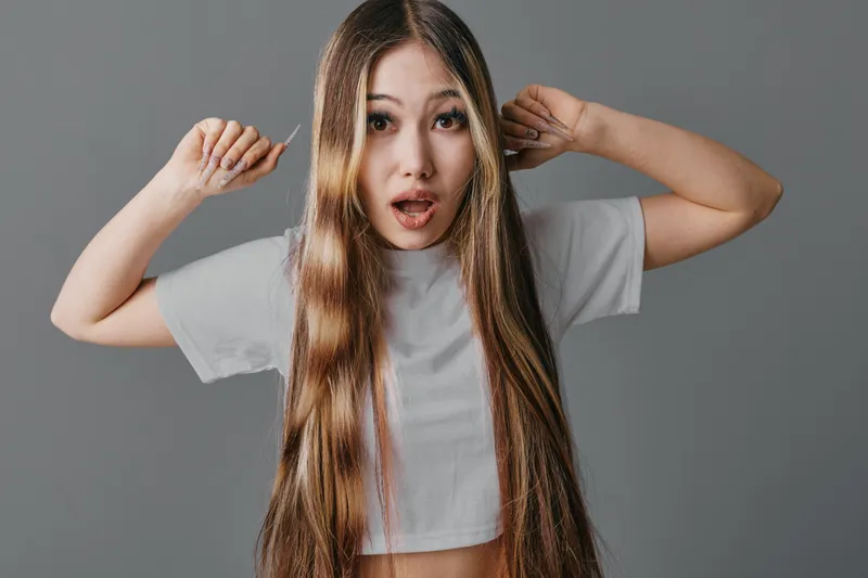 Young woman with long hair wearing casual white t shirt looks surprised and raises hands against isolated gray background in studio portrait.