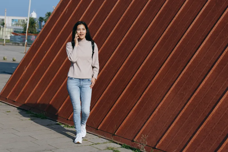 Casual outdoor scene with a woman talking on a phone beside a tall slanted wall, jeans and sweater, urban street setting, daytime light, relaxed candid lifestyle moment