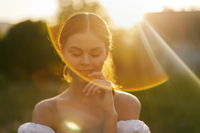 Beautiful young woman in a white dress softly smiling with a serene expression during sunset, capturing a moment of peaceful reflection in nature