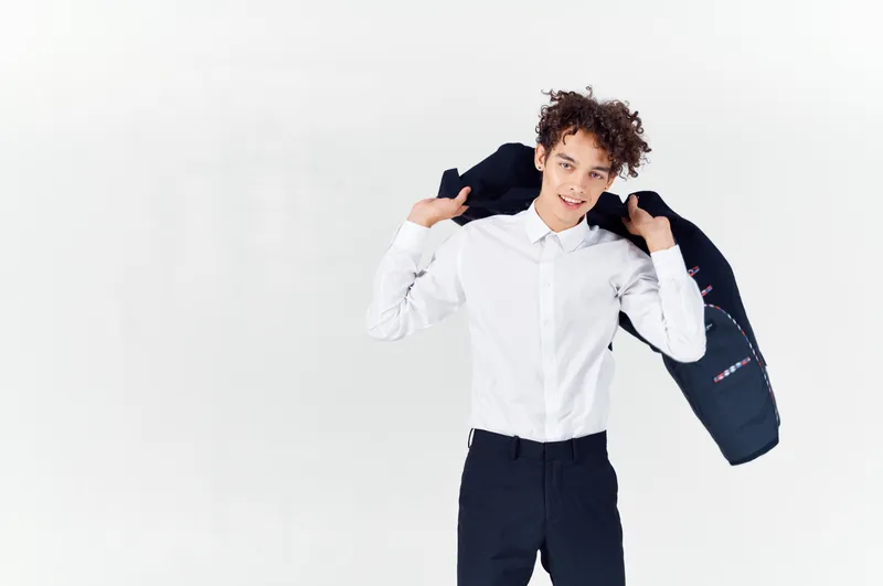 Подросток в классическом костюме держит в руке пиджак на светлом фоне, кадр обрезан до брюк