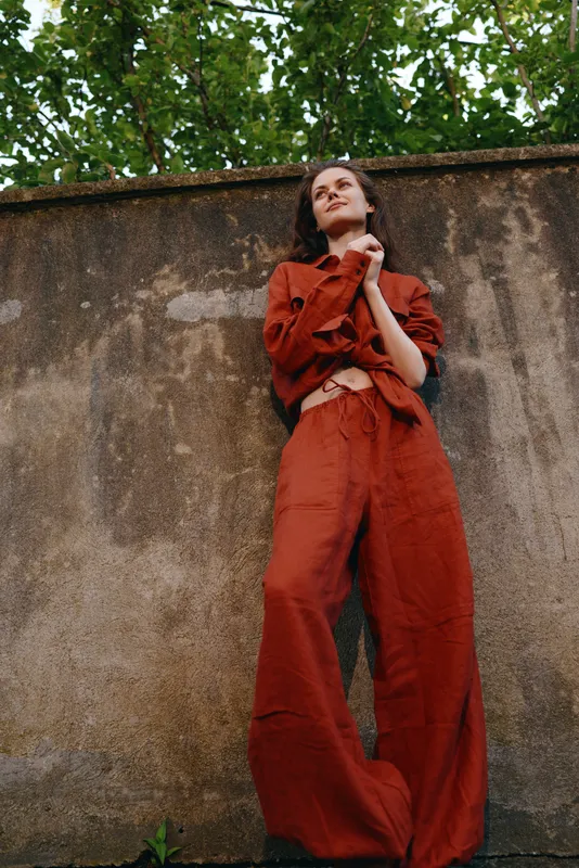 Confident woman outdoors wearing a rust jumpsuit, relaxed pose, sunlit moment, textured wall backdrop, lush greenery overhead, natural colors, casual lifestyle portrait with soft daylight mood