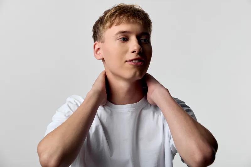 Young man in a white t shirt adjusts his neck in a studio setting, thoughtful expression, clean background, casual style, minimal lighting.