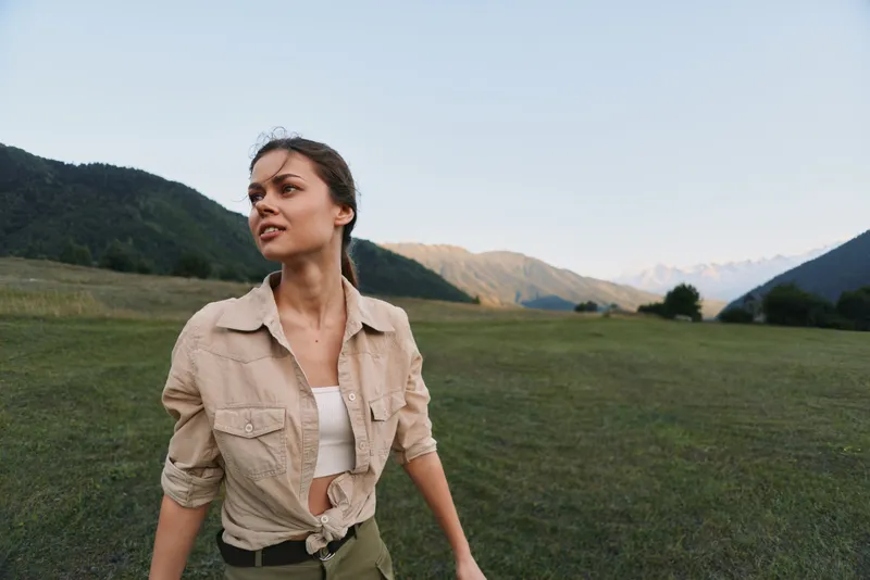 A young woman stands in a green meadow beneath distant mountains, wearing a beige shirt over a white top, looking upward with a calm, adventurous expression.