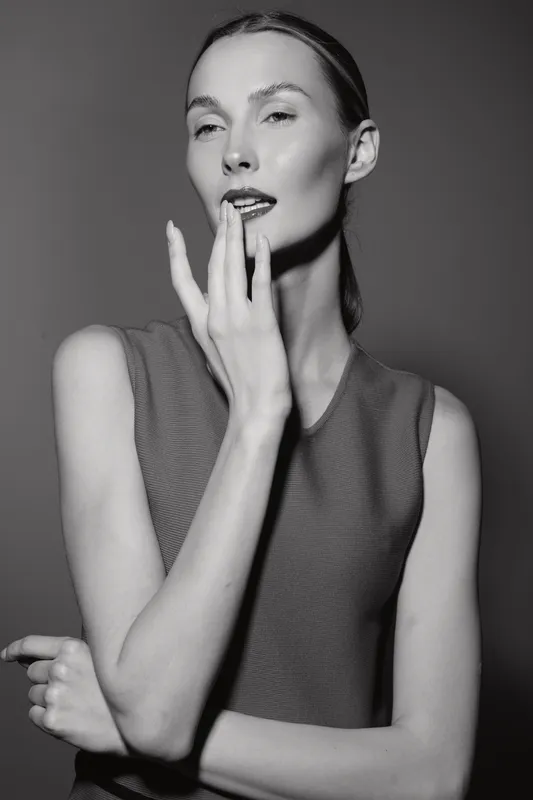 Slim Caucasian woman with fair skin and light hair poses with hand near lips in black and white studio portrait.