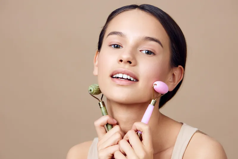 Young female model showcasing skincare tools, smiling with jade and rose quartz facial rollers against a soft beige background, conveying beauty and wellness