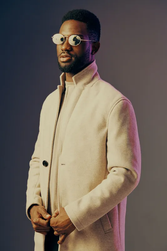 Stylish black male model in a beige coat and stylish sunglasses, showcasing modern fashion and confidence against a colorful studio background