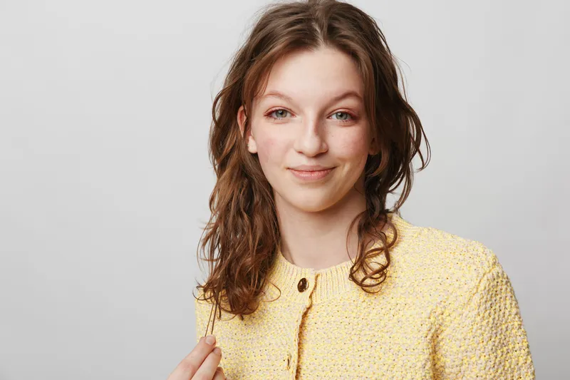 Young woman with curly brown hair wearing yellow knitted sweater smiles gently on grey background. Portrait of casual style, natural beauty, and confident female with relaxed expression.