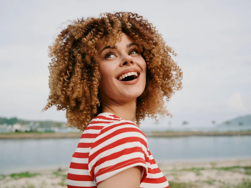 Joyful portrait of a young woman with curly hair wearing a red and white striped shirt, smiling up at the sky against a seaside background, with warm sunlight highlighting natural texture and