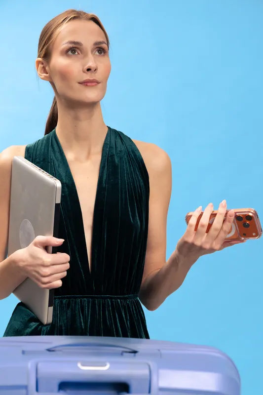 Young woman in green dress holds laptop and smartphone while standing behind blue suitcase. Studio portrait on blue background. Concept of travel, work, digital nomad, and business trip.