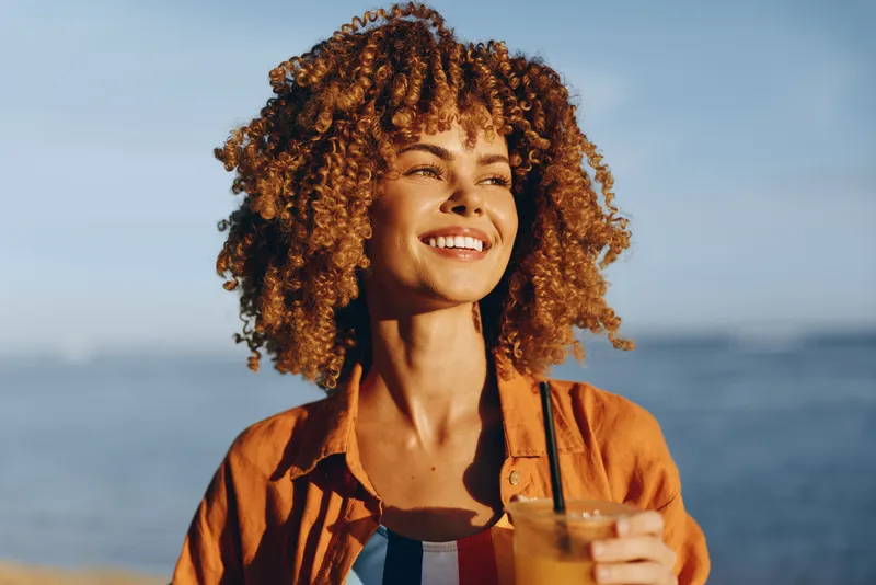 Young woman with curly hair smiles at the seaside, enjoying a sunny day with an orange drink, wearing an orange shirt, relaxed and joyful by the coast, her hair glowing in warm light as waves roll