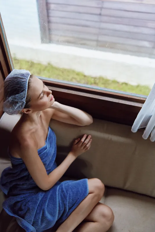 Relaxed woman wrapped in blue towel with shower cap sits thoughtfully by window, enjoying calm indoor moment and soft natural light in cozy spa-like setting.