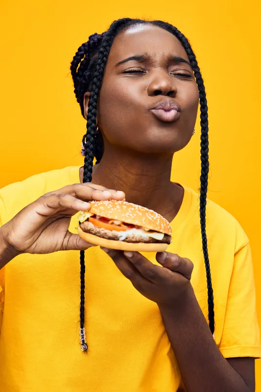 Happy young woman enjoying a burger with a playful expression against a vibrant yellow background, showcasing comfort food and joy in a trendy, colorful style