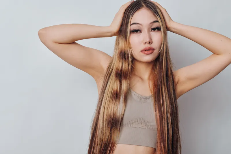 Young woman with long straight hair posing with hands on head against neutral background, wearing sleeveless top for fashion and beauty concept.