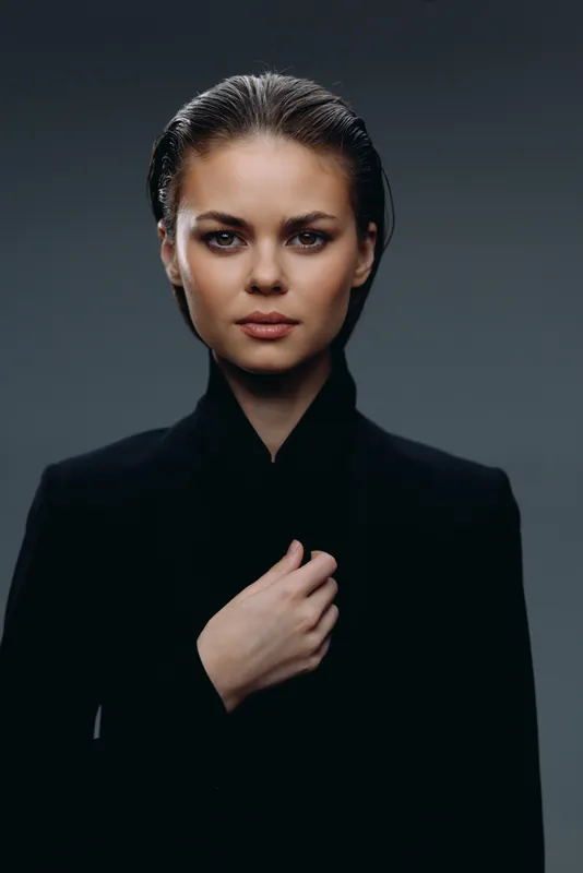 serious woman in black coat with hand on chest against dark background expressing calm confidence