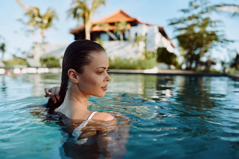 Woman in a pool with a calm expression, sunlit water, and a tropical resort backdrop, conveying relaxation and a casual vacation mood in natural colors