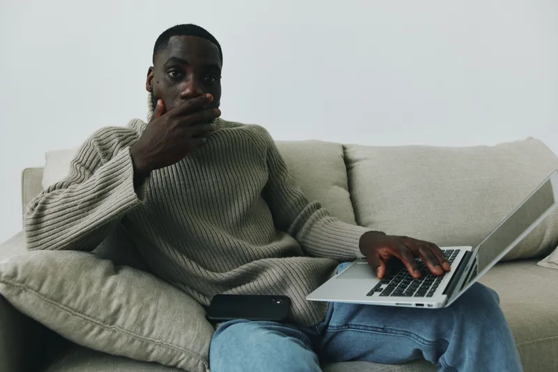 Thoughtful young African American man working on a laptop in a cozy living room, wearing a knitted sweater, expressing concern or contemplation