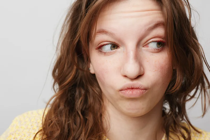 Close-up of a young woman with curly hair making a funny face, looking sideways. Casual portrait on a light background with natural expression and soft lighting.