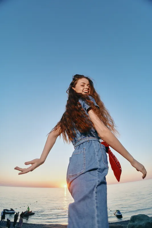 Lifestyle woman in modern boho-western denim street style smiling outdoors near ocean at sunset with film color effect and relaxed joyful mood.