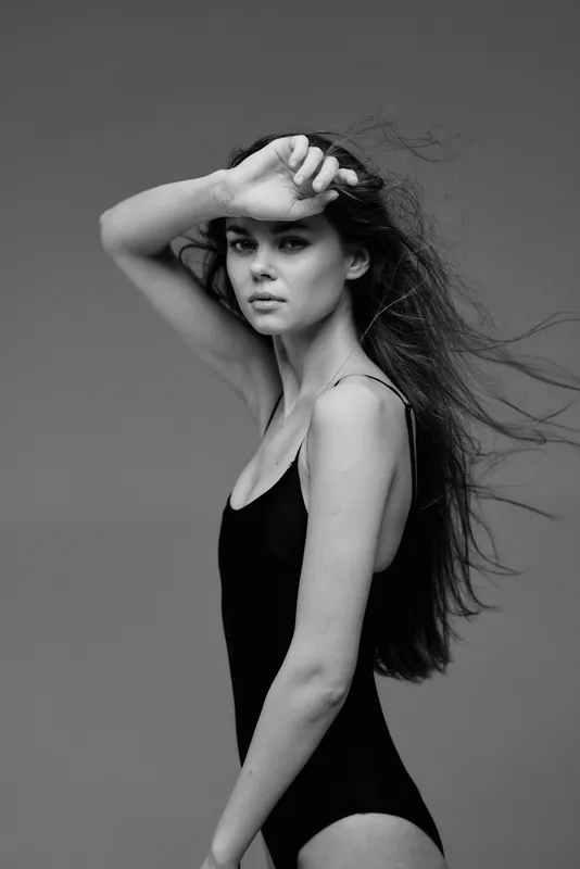 Black and white portrait of a young woman with long hair wearing a black bodysuit posing with hand on forehead.