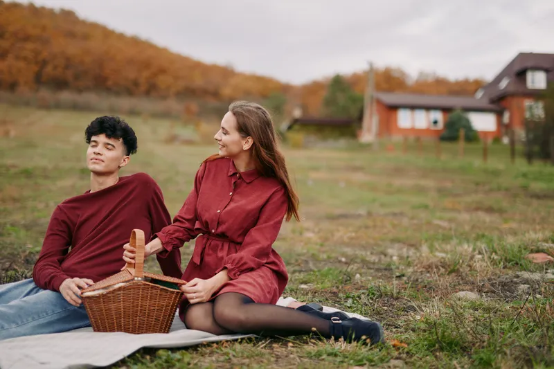 Two young adults share a simple picnic on a fall field, a candid moment that conveys authenticity and credibility as they relax on a blanket near rustic houses.