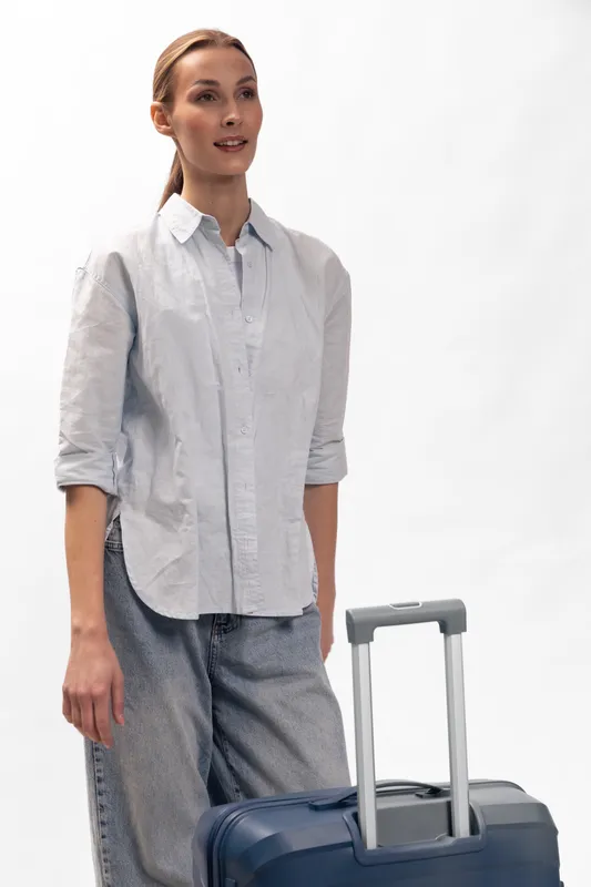 Young woman in casual clothes stands with blue suitcase on white background, looking forward. Concept of travel, vacation, tourism, and business trip with relaxed mood.