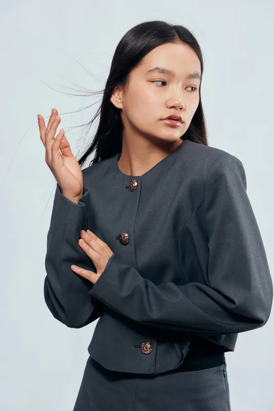 Young woman with long dark hair wearing gray outfit with buttons posing sideways on light background. Model shows subtle expression with hand raised near face and medium skin tone.