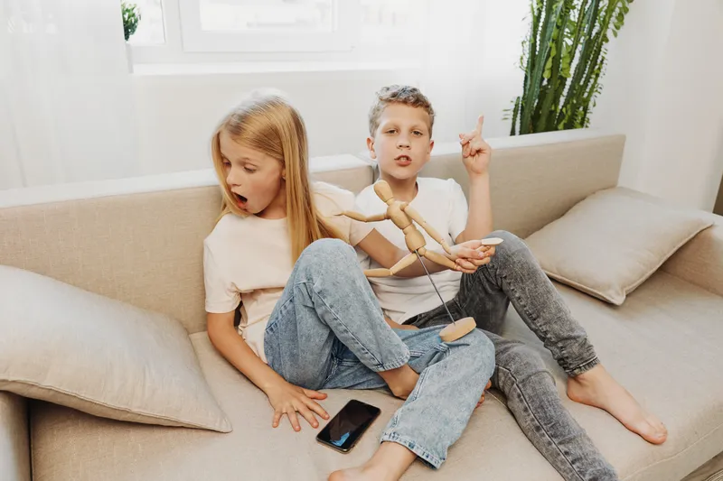 Two children sitting on a beige sofa playing with a wooden mannequin figure, surprised girl looking at the toy, casual home interior with natural light and green plant in background.