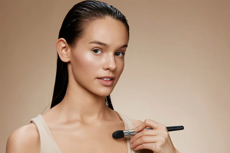 Young woman applying makeup with a brush, showcasing natural beauty and confidence against a soft beige background Ideal for beauty and skincare concepts