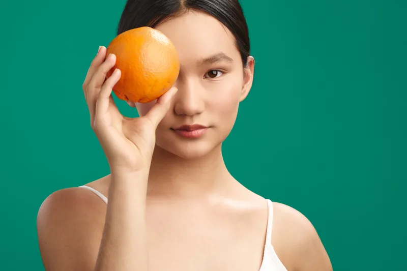 Asian woman holds an orange beside her eye in a vibrant studio portrait, against a bright green backdrop. The composition conveys playfulness, curiosity, and a calm, modern sense of style.