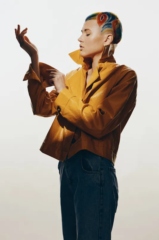Woman with colorful hair and stylish jacket posing gracefully against white background, showcasing modern fashion and artistic vibe in a minimalistic studio setting.