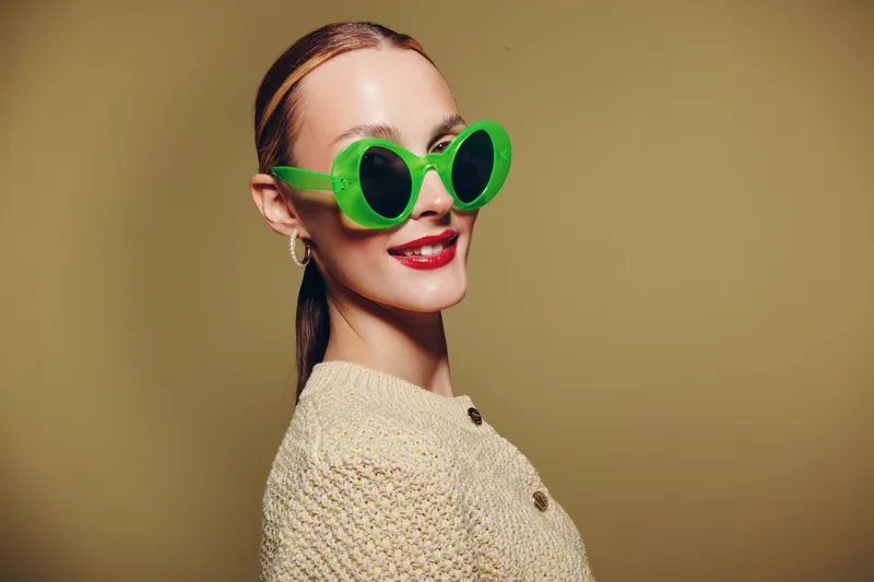 Smiling woman with green round sunglasses and red lipstick poses in beige knitted sweater on neutral background. Concept of fashion, style, accessories, and confident attitude.