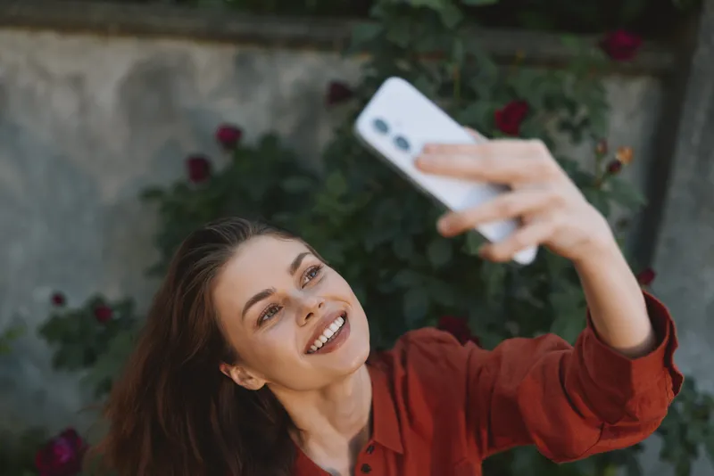 Woman lifestyle selfie outdoors in a garden, smiling brightly while holding a phone, wearing red casual attire, capturing a joyful portrait in natural daylight