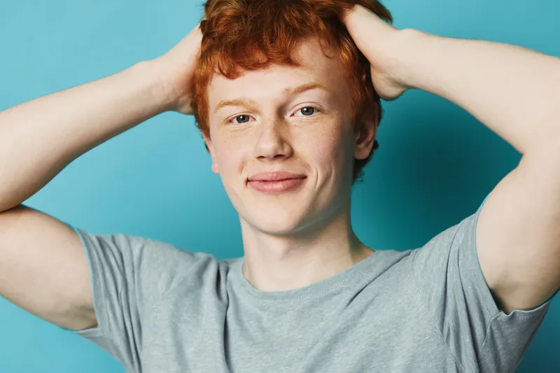 Young man with red hair wearing a gray t shirt smiling and holding hands behind head against blue background. Portrait of confident male with casual pose and natural expression.
