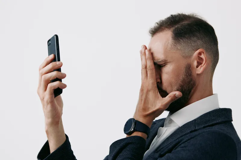 A professional man in business attire stands against a vivid isolated colored backdrop, conveying stress and focus as he checks his smartphone during a moment of doubt.