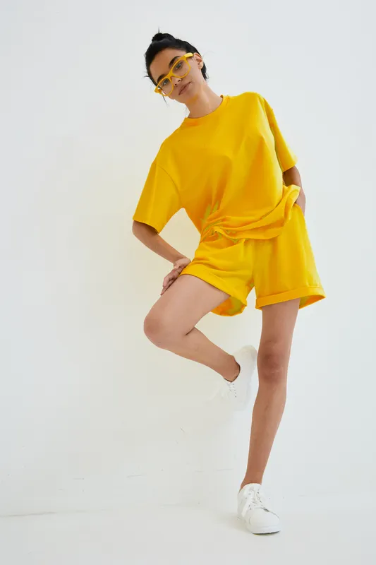 young woman in yellow outfit posing confidently against a white background, wearing sunglasses and white sneakers, modern casual fashion style