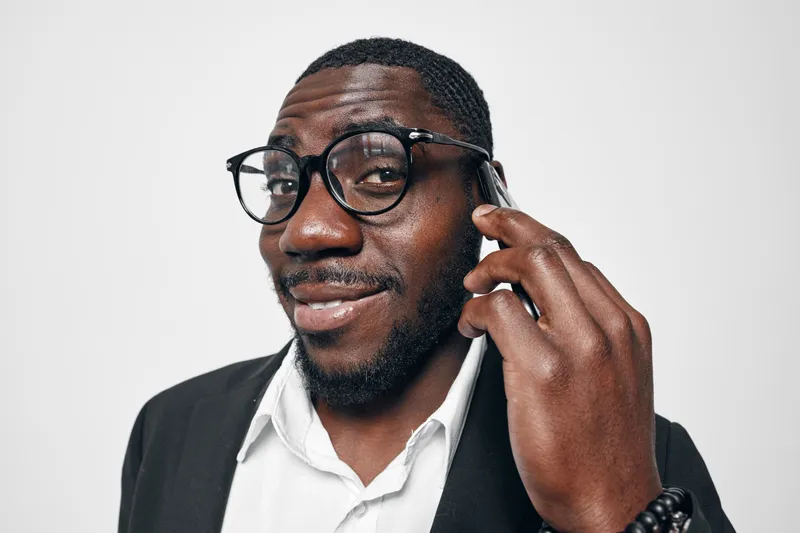 African American man with dark skin wearing glasses and black suit talking on smartphone with confident smile isolated on white background. Businessman, communication and technology concept.