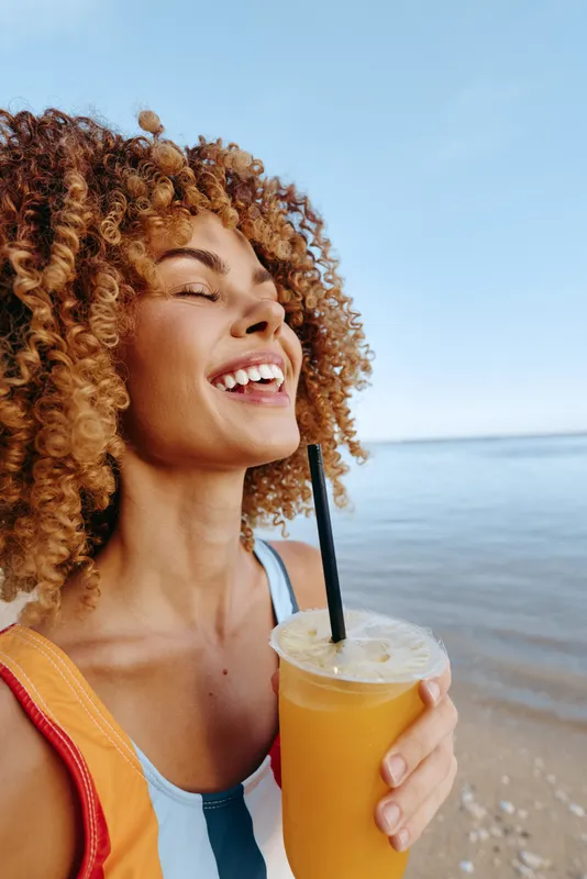 Young woman with curly hair enjoys a sunny beach moment, sipping orange juice with a bright smile and closed eyes as a light breeze lifts her hair, while the calm sea stretches behind.