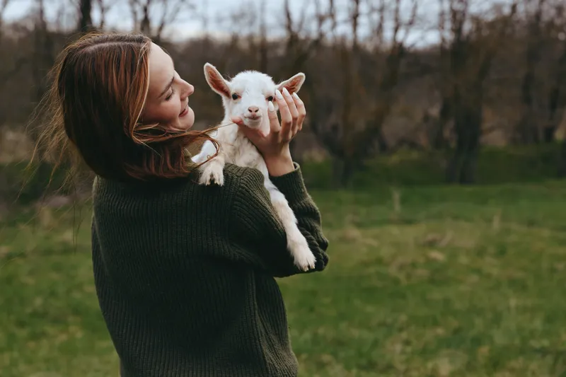 A woman in a dark sweater holds a fluffy goat kid in a grassy field, smiling softly. A lifestyle moment highlighting gentle bonding with animals and rural living.