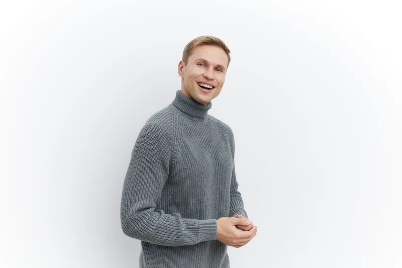 Happy smiling man wearing cozy gray turtleneck sweater standing against plain white background. Caucasian male adult with cheerful expression, casual style, indoor studio shot. People lifestyle concep