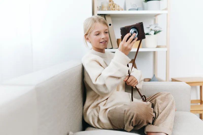 A young girl sits on a light sofa, holding a camera with a strap, capturing a moment indoors in a cozy, soft lit living space.