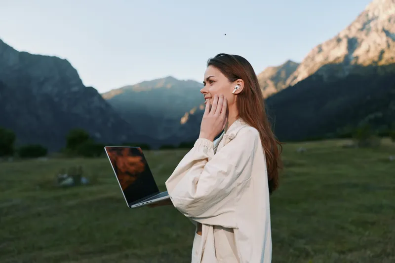 A thoughtful woman stands in a scenic outdoor meadow with a laptop, gazing into the distance as rugged mountains rise behind her, embodying focus and creative work in nature.