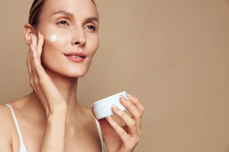 Young woman applies moisturizing cream on face holds jar with skincare product has smooth skin natural makeup and beige background beauty and health concept