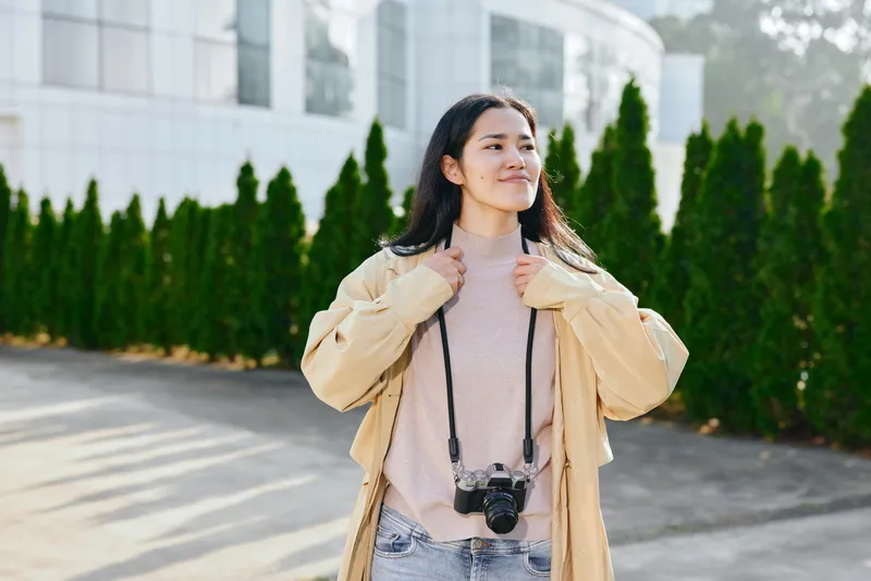 Woman with camera hanging around neck outdoors wearing beige jacket and pink sweater, calm smile, modern campus style, trees and glass building backdrop, candid fashion and travel vibe