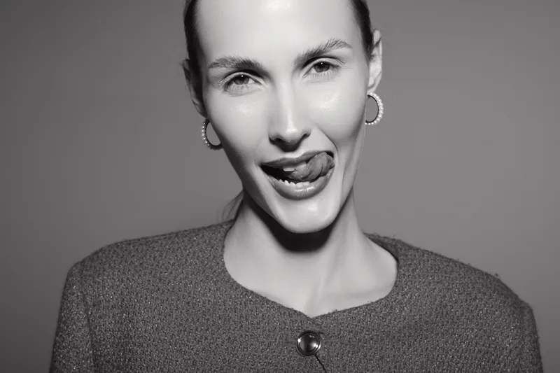 Close-up of a fair skinned Caucasian woman with slicked hair making a playful face, tongue out, wearing hoop earrings and textured sweater in monochrome studio lighting.
