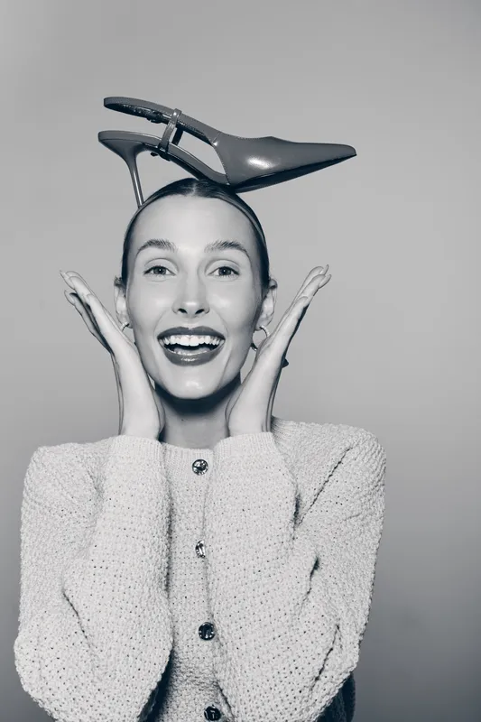 Happy caucasian woman with fair skin holds high heel shoe on head smiling excitedly wearing knitted sweater on grey background.