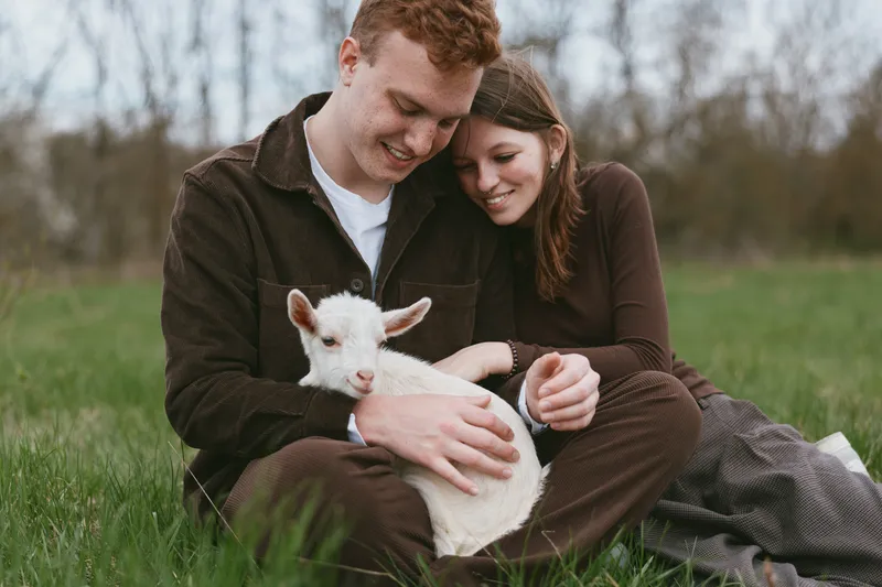 Couple outdoors with a goat kid in a meadow, enjoying affection and happiness under sunlight, a nature driven rustic lifestyle moment in a pastoral setting, candid and warm scene