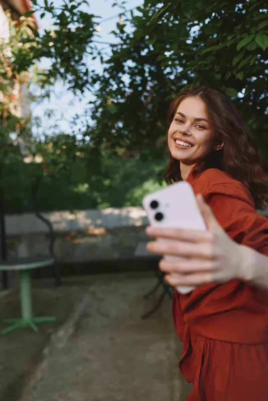 Smiling young woman in an orange dress takes a casual selfie in a sunlit garden, enjoying a carefree moment amid green leaves and a warm breeze, her natural pose conveying spontaneity