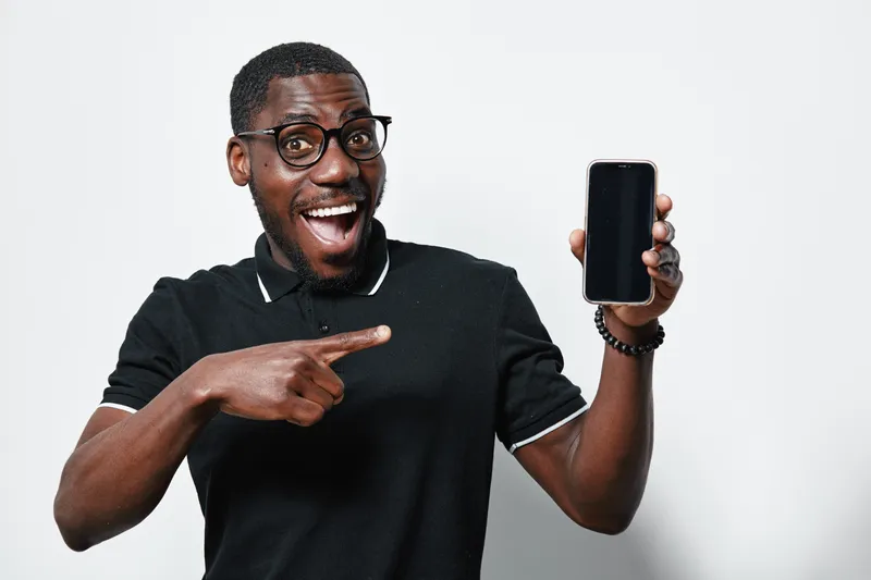 African American man with dark skin and glasses smiling and pointing at smartphone screen in black polo shirt on white background. Excited male showing mobile phone with blank display.