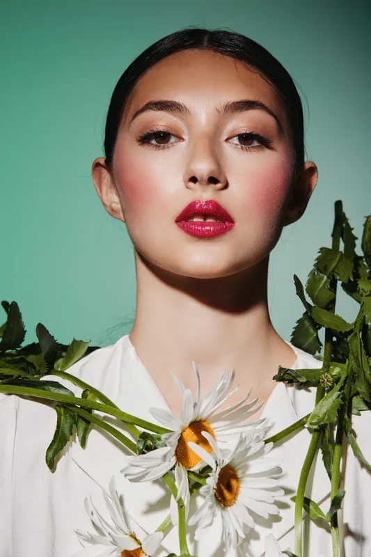 Close-up of a young woman with glossy red lips and soft pink blush wearing daisies over pale outfit. Creative beauty fashion shot on pastel green background.