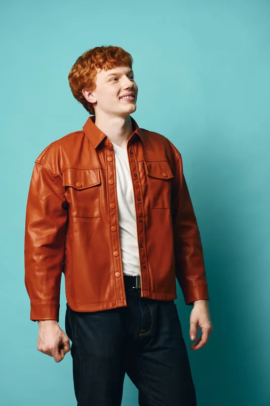 Young man with red hair wearing brown leather jacket and white t shirt standing against blue background looking aside with slight smile and casual posture.